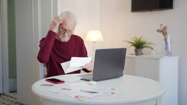An elderly man pays taxes via the Internet. enough money He scratches his head thoughtfully and examines bills The pensioner is engaged in paper work. He sends bills and tax receipts through a laptop. He looks confused. High quality 4k footage Retirement stock pictures, royalty-free photos & images
