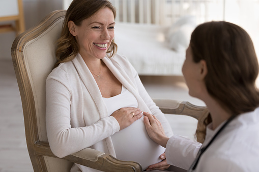 Obstetrician doctor visiting happy pregnant patient woman at home, touching belly, examining abdomen, feeling baby kick, talking to expecting mom, laughing. Prenatal care concept