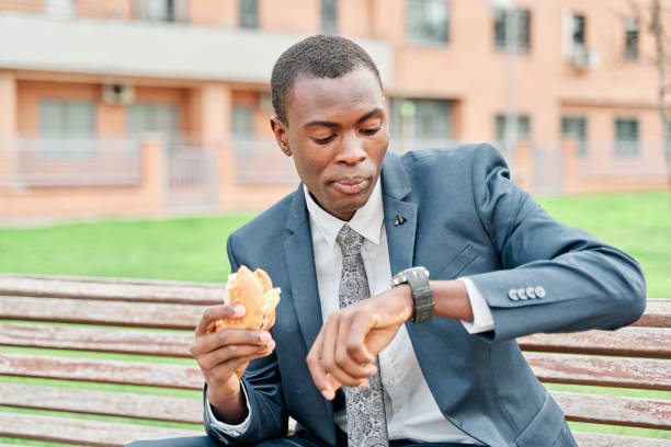 african-american businessman eating a hamburger, watches wristwatch sitting on a bench in the park. - eating sandwich emotional stress food imagens e fotografias de stock