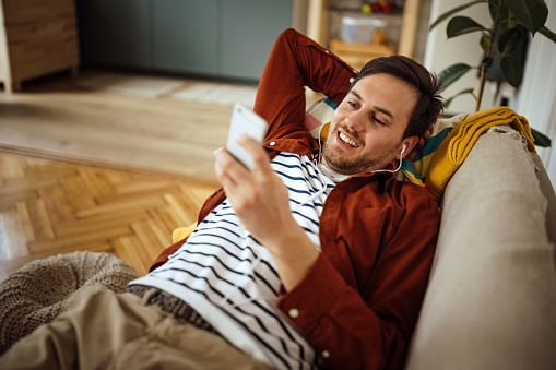 High angle view of smiling young man listening music through headphones and using smart phone while relaxing on sofa at home