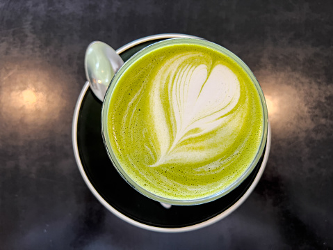 Horizontal high angle closeup photo of a glass of vibrant green Matcha latte with a heart shaped froth art on a black saucer with a teaspoon.