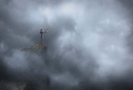 A man stands at the edge of a steep cliff as he looks through a spyglass while attempting to establish his bearings in the thick fog that surrounds him.