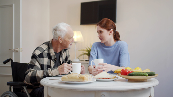 A young woman and an elderly disabled person in a wheelchair sit at a table and drink tea. Fruit and pastries are on the table. People talk nicely. High quality 4k footage