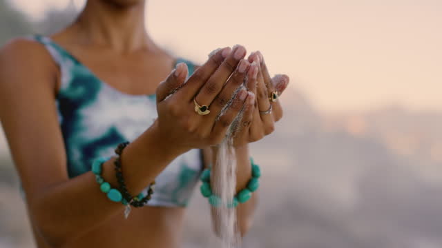 4k video footage of a woman letting sand slip through her fingers while doing yoga on the beach at sunset