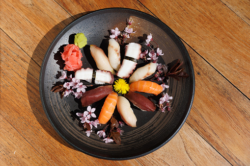 Plate of various nigiri pieces served with wasabi and ginger on wooden table, decorated with cherry blossom and dandalion