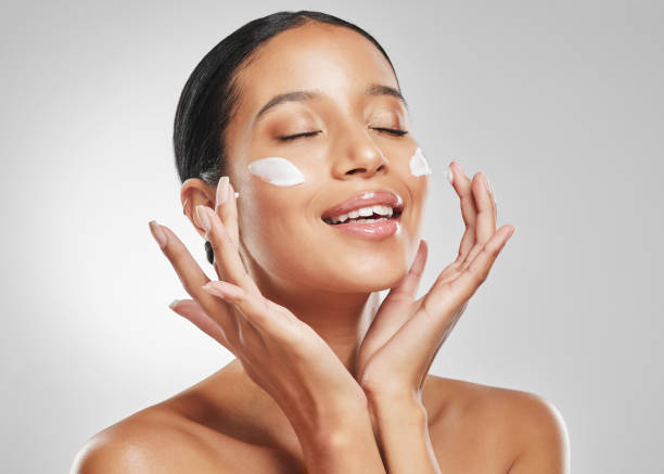 studio shot of an attractive young woman posing with moisturiser on her face against a grey background - applying imagens e fotografias de stock