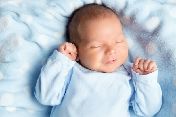 bebé recién nacido durmiendo sonriendo. lindo niño en traje de cuerpo envolvente. niño recién nacido sonríe con ropa azul acostado en la manta. cara de niño pequeño retrato de cerca sosteniendo las manos primero - sleeping baby fotografías e imágenes de stock