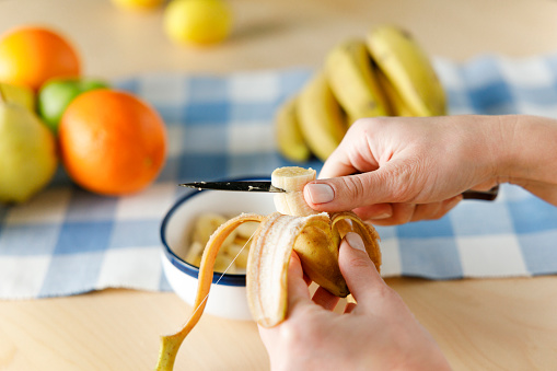 Woman sitting by the table at home cutting banana. She is preparing fruit salad at home