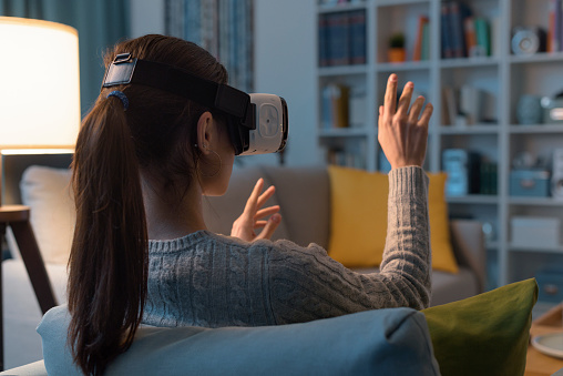 Young woman sitting on the sofa at home and wearing a VR headset, she is interacting with virtual reality