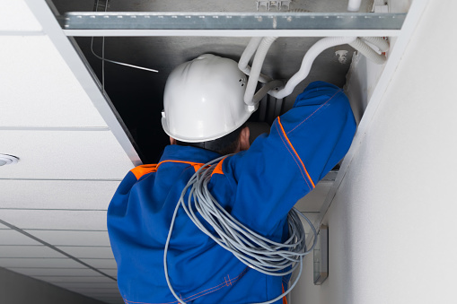 foreman in blue uniform and white hard hat laying wiring for a Wi-Fi router in a hidden false ceiling system, close-up