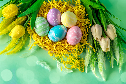 Multi colored hand-painted Easter eggs in a basket with some tulips flowers. Green defocused lights background with beautiful bokeh. Space for copy.