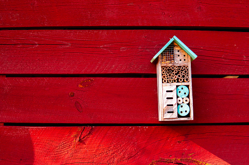 A design insect hotel hanging on a red wall outside in the daytime sun.
