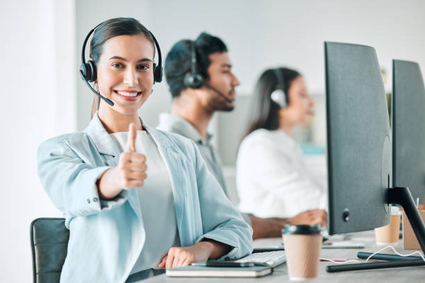 retrato de uma jovem agente de call center mostrando polegares para cima enquanto trabalhava em um escritório com seus colegas ao fundo - female customer service representative looking at camera technology - fotografias e filmes do acervo