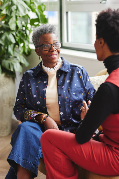 ritratto di due donne d'affari nere sedute sul divano e che hanno una conversazione rilassata, donna anziana con occhiali che ascoltano e sorridono - relazione daffari foto e immagini stock