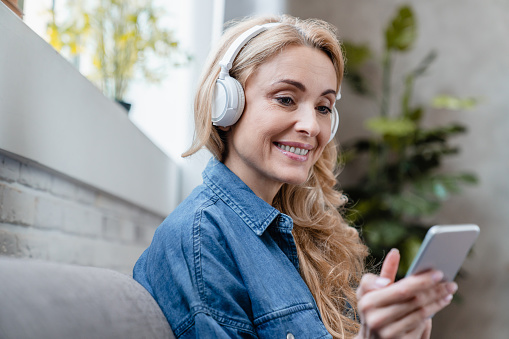 Mature middle-aged caucasian woman enjoying music in mobile application and headphones, choosing radio podcast e-book online relaxing on the couch sofa at home