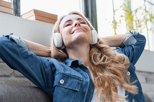 Music phone application. Relaxed happy serene mature middle-aged caucasian woman listening to the music in headphones earphones, meditating, radio podcast e-book online at home