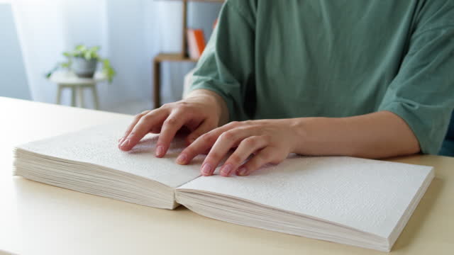 Blind young man reading braille book using his fingers, sitting in living room, poorly seeing person learning to read, home education for people with disabilities, touching letters on sheet of paper