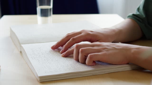 Blind person reading braille book, touching letters on sheet of paper using his fingers, sitting at desk, poorly seeing man or woman learning to read, home education for people with disabilities