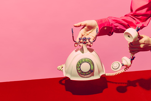 White telephone, Pop art photography. Retro objects, gadgets. Female hand holding handset of vintage phone isolated on pink and red background. Vintage, retro 80s, 70s style. Complementary colors.
