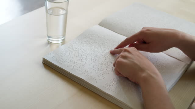 Blind man reading braille book, touching letters on sheet of paper using his fingers, sitting at desk, poorly seeing person learning to read, concept of home education for people with disabilities