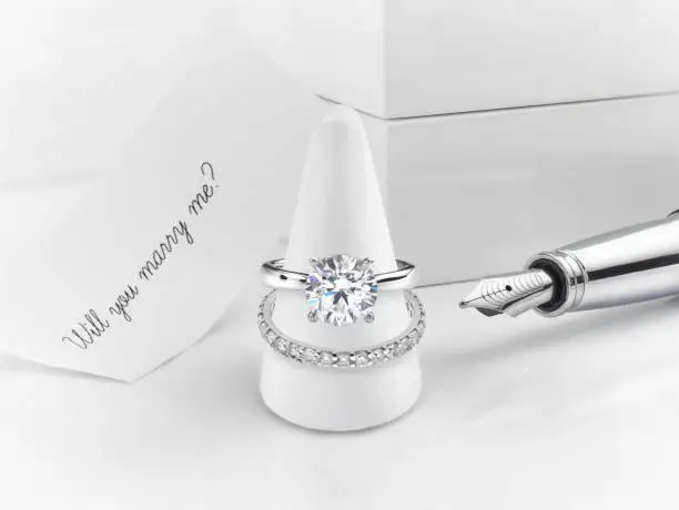 Solitaire engagement ring with diamond wedding ring on display cone. Accompanied by proposal note. Will you marry me, alongside fountain pen.