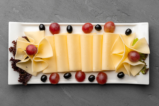 Cheese platter with grapes and black olives on gray background, close up