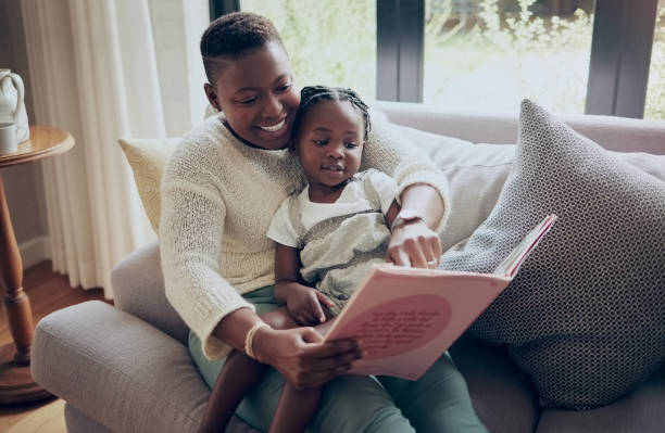 shot of a mother reading a book with her daughter at home - family reading african descent book imagens e fotografias de stock