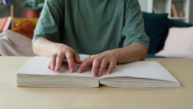Blindman reading braille book using his fingers, sitting in living room, poorly seeing person learning to read, home education for people with disabilities, touching letters on sheet of paper