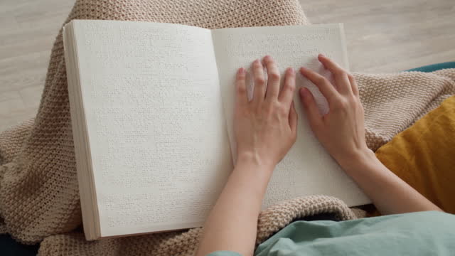 Reading braille book sitting on sofa, blind person touching letters on sheet of paper using his fingers, poorly seeing woman learning to read, concept of home education for people with disabilities