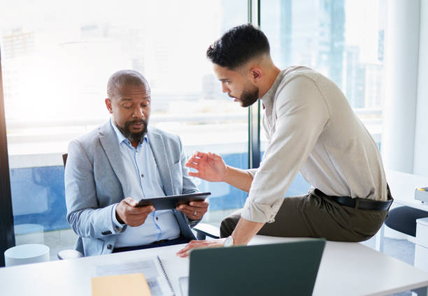 istock Shot of two businessmen sitting in the office together and having a discussion while using a digital tablet 1389488096