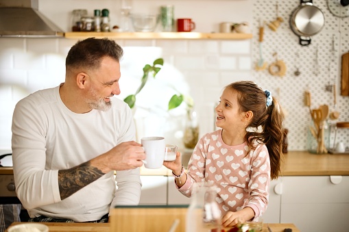 A loving dad teaches his daughter to cook. Dad and little girl prepare pancakes for breakfast. Father and daughter in pyjamas sitting in the kitchen in the morning