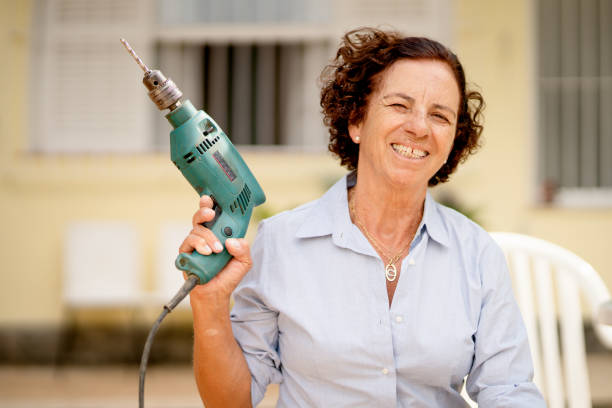 Smiling mature woman holding a drill while doing some diy at home Portrait of a smiling mature woman holding a drill while doing some diy in her back yard at home in summer holding drill stock pictures, royalty-free photos & images