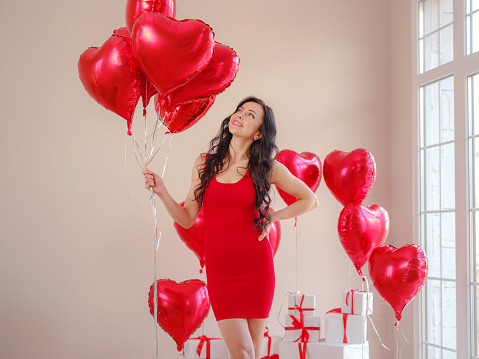 Beautiful happy young woman in red dress. holiday party. Joyful model posing with red heart shaped balloons, having fun, celebrating Valentine's Day. Symbol of love
