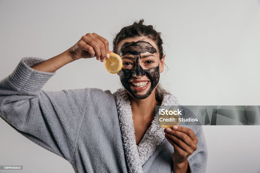 I care for my skin Portrait of a beautiful young adult woman using beauty treatment, a carbon face mask, while holding two lemons, rubbing her skin with them, hydrating her skin and having a soft smile, while  standing isolated on a gray background, smiling at the camera She is wearing a bathrobe, facing the camera Merchandise Stock Photo