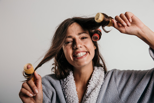 Close up image of a smiling young woman with rollers in her hair, wearing a bath robe, preparing for the day. Shot against studio background.