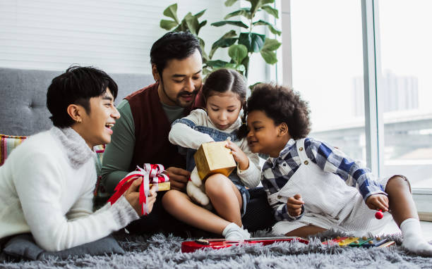 couple male gay taking care, giving gift to adopted children who are little diverse caucasian and african girl and boy with happiness to celeblate birthday in living room at home. lgbt, kids concept. - child little boys male caucasian imagens e fotografias de stock