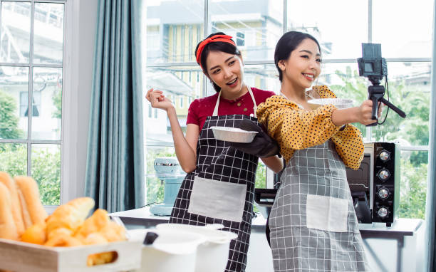 istock Two beautiful Asian women cooking food, making bread for breakfast and bakery while using tablet, streaming live video clip or taking selfie photo together with happiness and fun in kitchen at home. 1389507197
