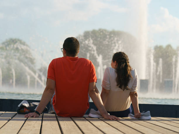 Heterosexual couple of young woman and man resting in front of fountain in the city public park in day Moscow, Russia - July 6, 2020: Heterosexual couple of young woman and man resting at the bench in the city public park. Time to rest in hot summer day. Youth lifestyle. Backs, rear view. moscow russia stock pictures, royalty-free photos & images