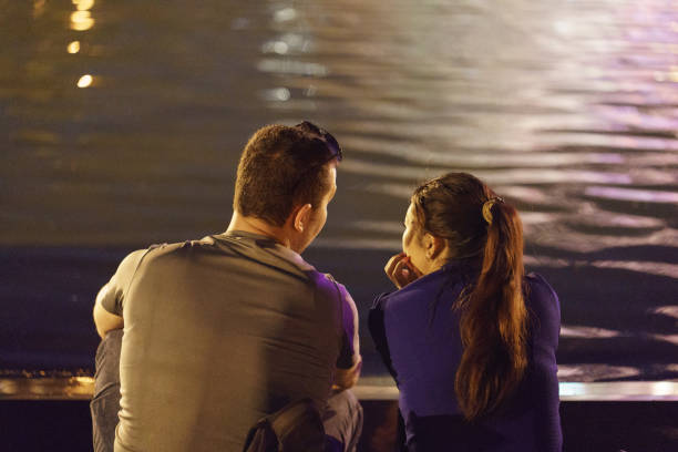 pareja heterosexual de mujer y hombre joven descansando frente a la fuente en el parque público de la ciudad por la noche - ponytail brown hair tourist women fotografías e imágenes de stock