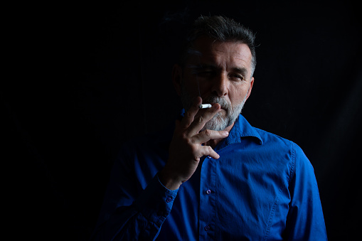 Handsome mature man smoking cigarette in dark room. dark and sullen shot of a mature man smoking over a black background. Man smoking cigarette on black background.