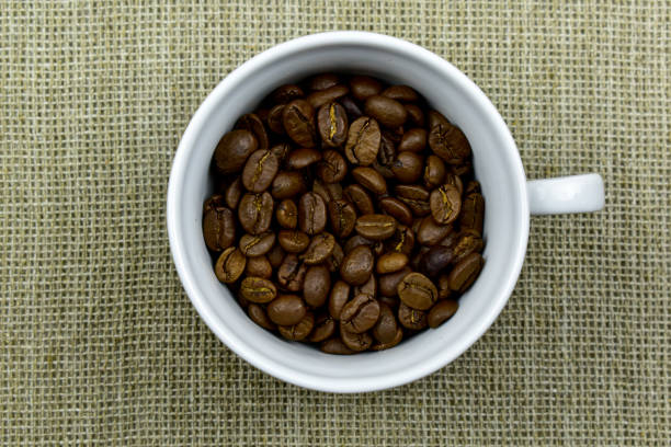 white cup filled with coffee beans, standing on burlap, top view, close-up