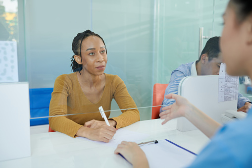 Black woman talking to nurse at clinic reception and filling form with personal data