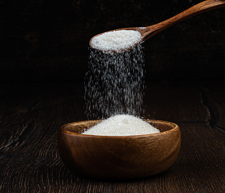 White granulated sugar is poured from a wooden spoon into a wooden bowl. Copy space.