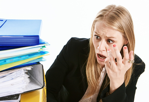 Young businesswoman or office worker gestures as she looks in horror at a stack of lever arch files full of paperwork.