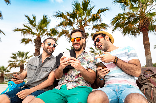 Multiracial group of friends using smartphone at seaside in Barcelona - Three young multiethnic men relaxing and having fun, smiling and talking together - Friendship and summer concepts