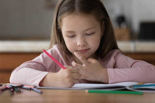 Sweet preschool girl kid using colored pencils, drawing in paper album, developing creative artistic skills. Little child engaged in home activities, sitting at kitchen table