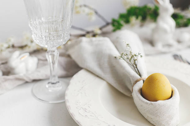 istock Stylish elegant Easter brunch table setting. Easter egg in bunny napkin on plate with cutlery, bunny, spring flowers on rustic white table. Modern Easter table arrangement 1389542579