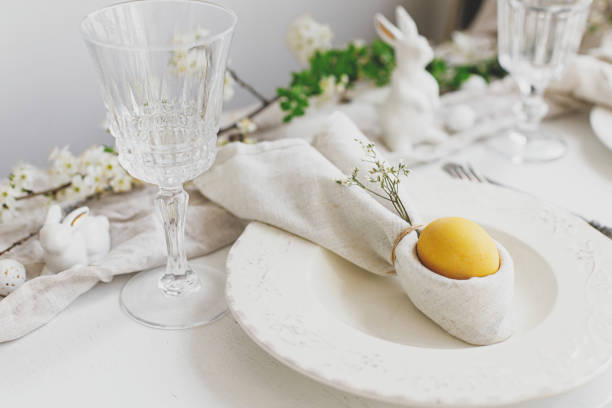 istock Stylish elegant Easter brunch table setting. Easter egg in bunny napkin on plate with cutlery, bunny, spring flowers on rustic white table. Modern Easter table arrangement 1389542613