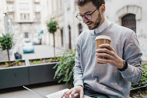 A young man is using a laptop and drinking coffee to go while sitting on the bench