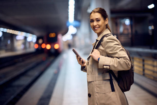 Happy woman using cell phone while waiting at subway platform and looking at camera.